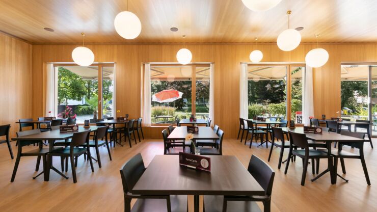 Plusieurs tables et chaises sont disposées à l'intérieur du restaurant, dont les murs et le sol sont boisés. À travers les larges fenêtres et baies vitrées, on voit la terrasse extérieure.