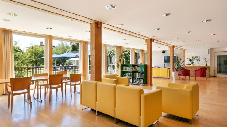 Des fauteuils jaunes et des tables ainsi que des chaises en bois clair sont disposés dans une vaste pièce lumineuse. À droite, on voit une grande baie vitrée donnant sur un jardin.