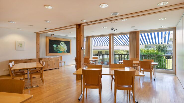Des tables rectangulaires en bois clair et des chaises sont disposées dans une salle revêtue d'un parquet en bois. En arrière-plan, on voit une baie vitrée.