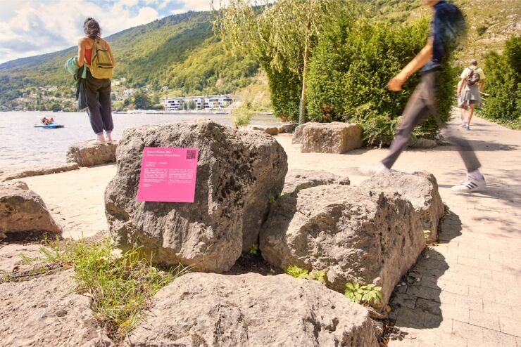 Eine rosafarbene Plakette mit Inschrift und QR-Code ist an einem Stein am Ufer des Bielersees angebracht worden. Um diesen herum gehen Leute Ihren Freizeitaktivit&auml;ten wie etwa spazieren und Boot fahren nach. 