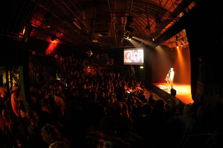 Ein grosser, dunkler Saal voller Menschen. Auf der rechten Seite befindet sich eine Bühne mit einem Mann darauf, der vor einem Mikrofon steht. Es gibt gelbe und rote Lichterspots, die vor allem auf den Mann auf der Bühne ausgerichtet sind. Daneben sieht man zudem eine Leinwandprojektion der Bühne. 