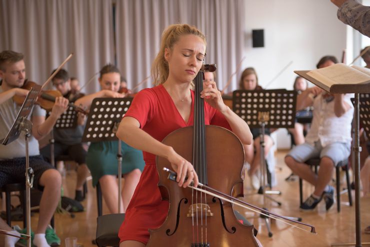 Dans une grande salle il y a beaucoup de jeunes qui jouent des instruments à cordes. Devant eux se trouvent les pupitres à musique. Au centre de l'attention se trouve une jeune femme blonde en robe rouge qui a les yeux fermés pendant qu'elle joue du violoncelle. 