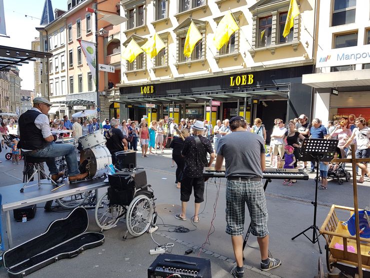 Auf einer Einkaufsstrasse haben sich viele Menschen um 4 Musiker herum versammelt. Diese bestehen aus einem Schlagzeug, einem E-Piano und 2 Gitarren. 