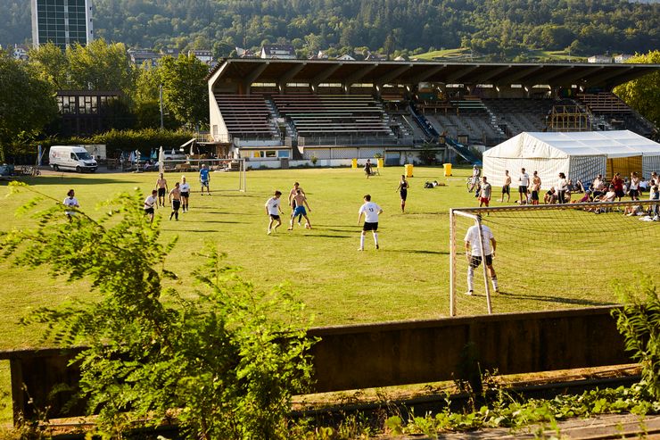 Auf einem grossen Rasen spielen Jugendliche miteinander Fussball. Im Hintergrund ist ausserdem ein Festzelt und eine Zuschauertribüne zu erkennen. 