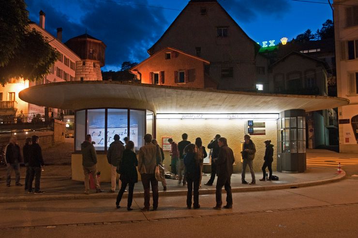 On dirait une vieille maison de tramway sur laquelle on peut lire les mots "Juraplatz". Devant lui se trouve un groupe de personnes qui se parlent devant une vitrine avec une photo. 