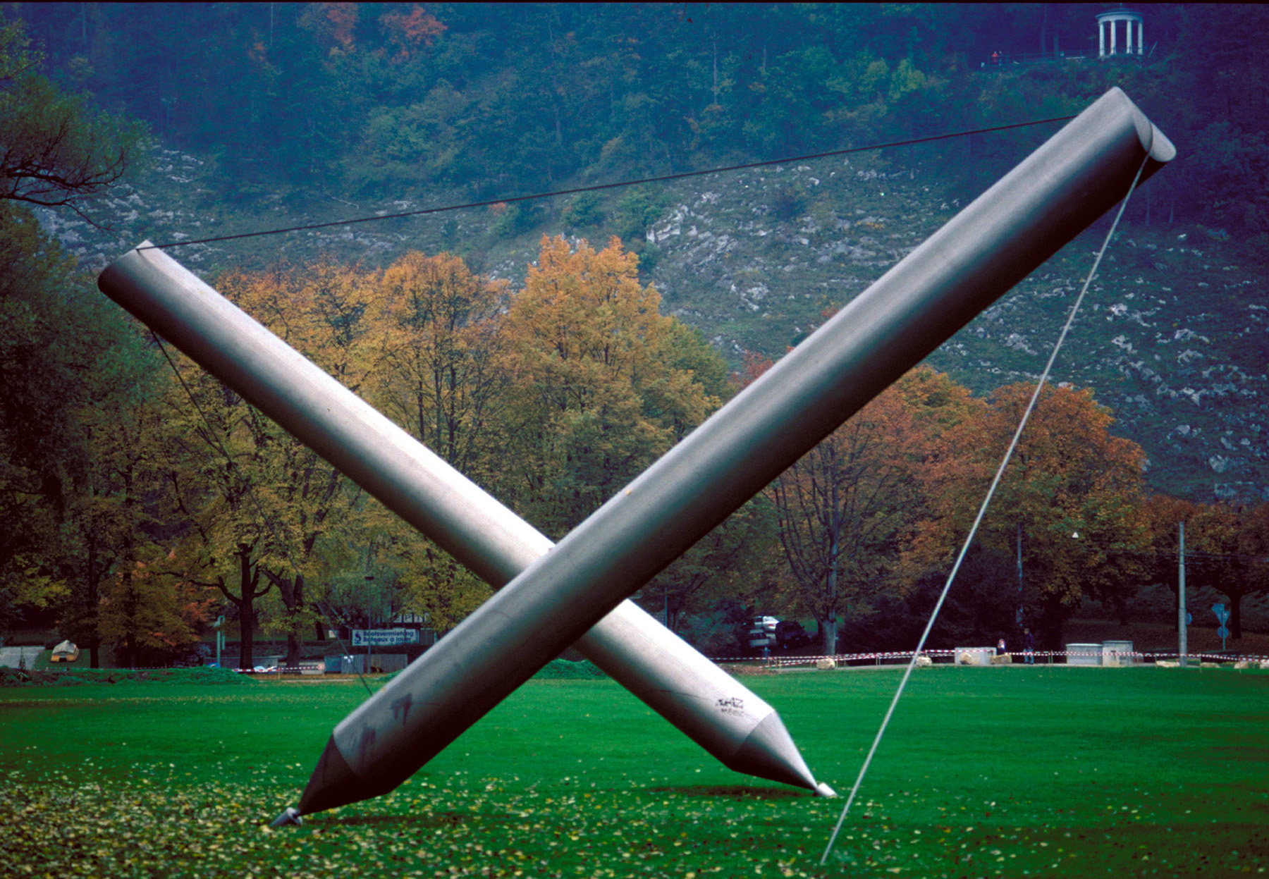 Une grande sculpture est installée sur une prairie. Il s'agit de deux stylos métalliques qui sont mises en place en étant tendues à l'aide d'un fil. Les pointes des stylos sont les points de contact avec le sol. 
