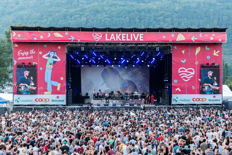 Une grande scène avec l'inscription "Lakelive" a été construite. Un groupe joue de la musique et 2 écrans diffusent la scène en direct. Devant elle, on peut voir une foule nombreuse. 
