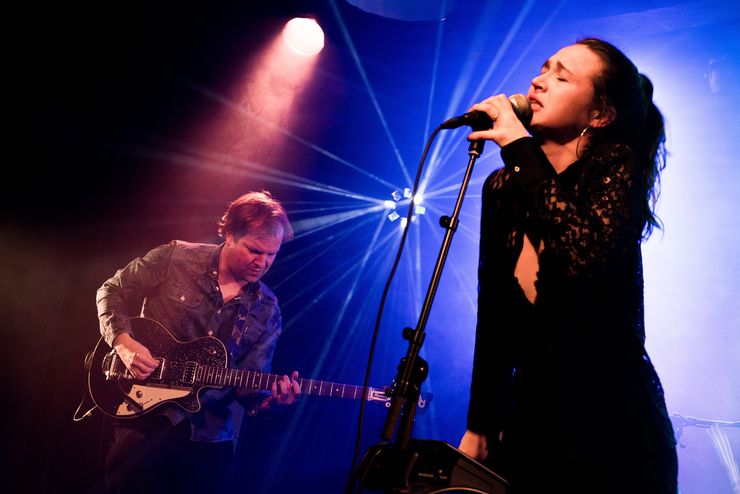 Auf einer Bühne, von einem Lichtspot beleuchtet machen zwei Leute Musik. Ein Mann an der Gitarre und eine Frau, die singt. 