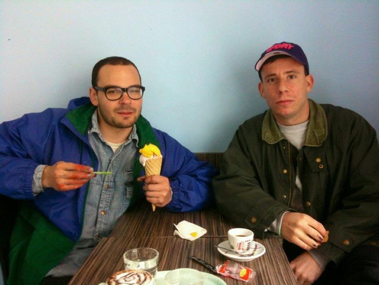 Portr&auml;t der Gewinner des Kulturpreises 2015, Linus Bill und Adrien Horni. Die beiden K&uuml;nstler sitzen an einem Tisch. Auf der linken Seite isst Linus Bill ein gelbes Eis. Er tr&auml;gt eine Brille, kurze Haare und eine blau-gr&uuml;ne Jacke. Adrien Horni trinkt Kaffee auf der rechten Seite. Er tr&auml;gt eine lila M&uuml;tze mit rosa Schrift und eine gr&uuml;ne Jacke.