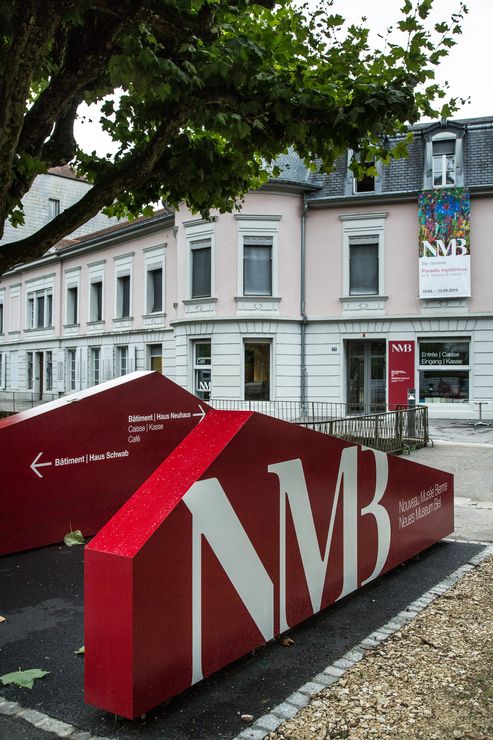 Au premier plan se trouve un panneau rouge moderne où on peut lire "Neues Museum Biel". Une flèche indique également "Haus Schwab" et l'autre "Haus Neuhaus", qui est déjà visible à l'arrière-plan. De plus, un arbre fait saillie dans l'image. 