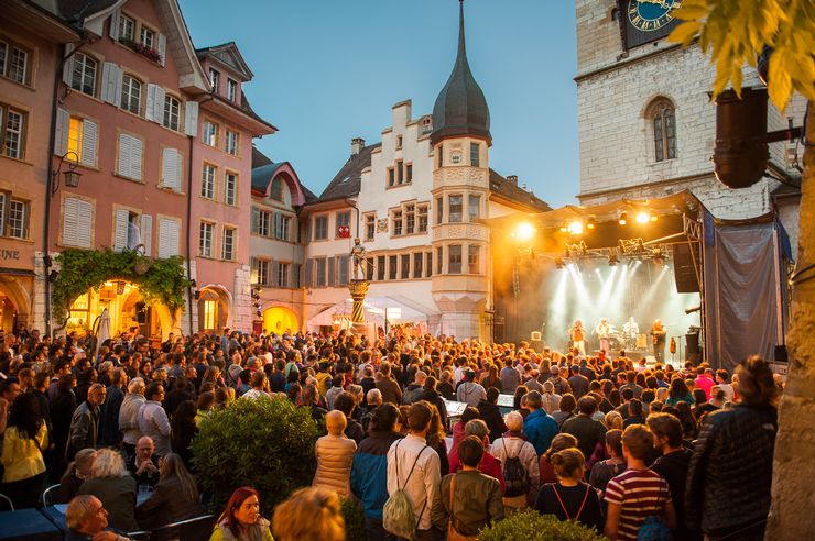 Auf einem Platz in der Altstadt von Biel ist eine grosse Bühne, auf der 4 Musiker performen und sehr viele Leute stehen davor, die der Musik lauschen. 