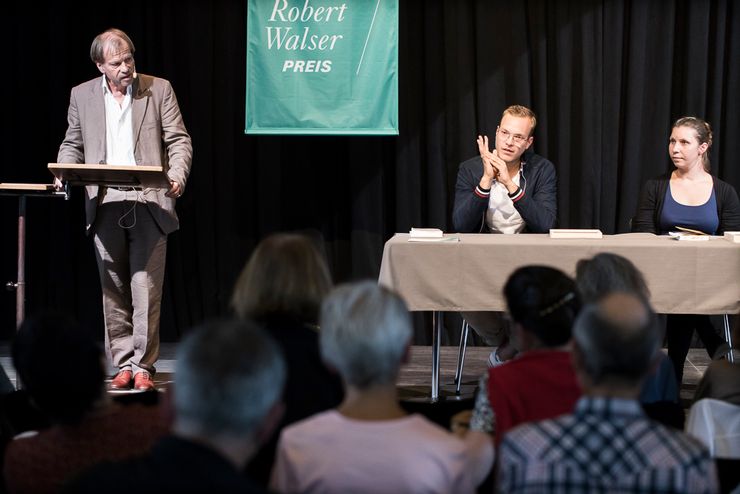 Ein Mann im Anzug mit einem Mikrofon ausgestattet steht an einem Rednerpult und spricht. Daneben eine Fahne mit der Aufschrift «Robert Walser Preis» und davor ein Mann und eine Frau an einem Tisch sitzend, mit Büchern vor sich ausgebreitet. Man erahnt auch ein grösseres Publikum davor. 