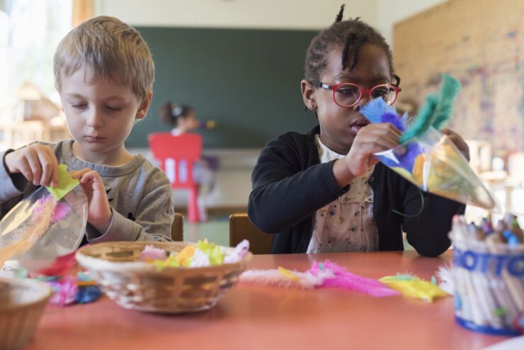 Zwei Kinder, ein M&auml;dchen und ein Junge, sitzen nebeneinander an einem Tisch und basteln mit farbigem Papier und Federn.