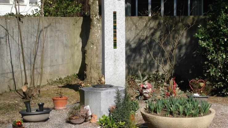 Es ist das Gemeinschaftsgrab vom Friedhof Bözingen zu sehen. In der Mitte steht eine ca. 5 Meter hohe Steinskulptur. Direkt davor befindet sich eine achteckige steinige, ca. 80 Zentimeter hohe Gemeinschaftsurne. Links und rechts davon haben Angehörige Blumenarrangements in Töpfen hingestellt.
