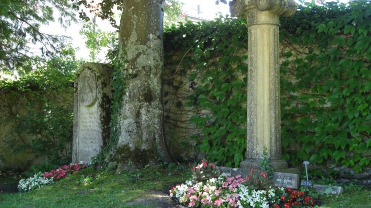 Es sind zwei alte Grabstellen auf dem Friedhof Madretsch zu sehen. Sie stehen direkt vor der Friedhofmauer, welche mit Reben überwachsen ist. Zwischen den Gräbern steht ein grosser Baum. Bei beiden Gräber zieren trotz ihres Alters verschiedene Blumen die Flächen vor den Grabsteinen.