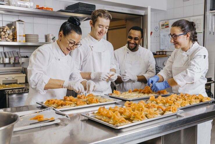 Vier lächelnde junge Köche und Köchinnen verschiedener Nationalitäten bereiten in einer Produktionsküche Gerichte zu.