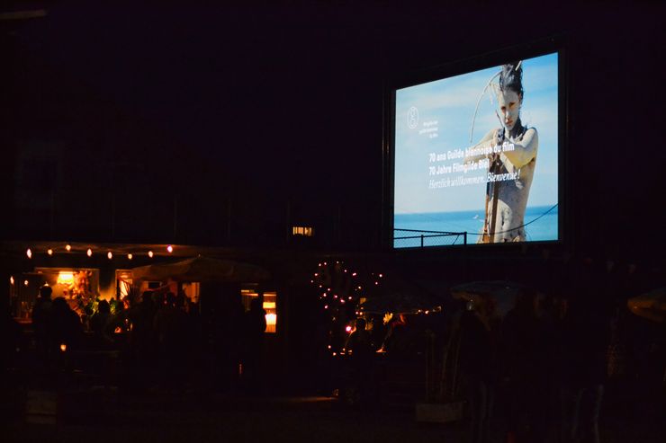 Il fait sombre et on est dehors. Sur le côté droit, on peut voir un grand écran de cinéma, sur lequel est écrit "70 ans Filmgilde Biel" et une femme peinte avec un arc à flèche est reconnaissable. 