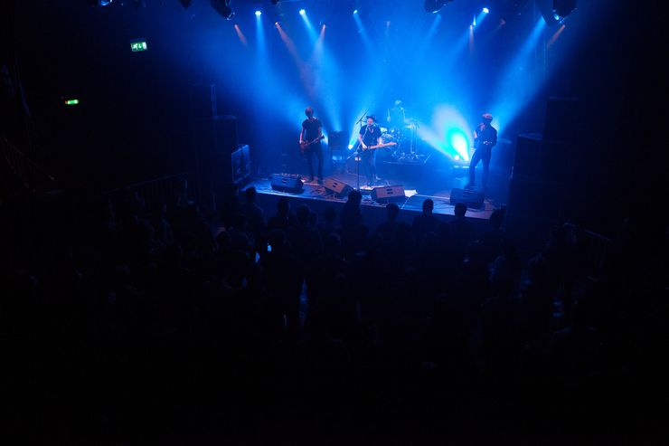 Eine Band bestehend aus 3 Gitarren und einem Schlagzeug spielt auf einer Musikbühne, sie werden blau beleuchtet, ansonsten ist es ziemlich dunkel und die Zuschauer sind nur zu erahnen. 