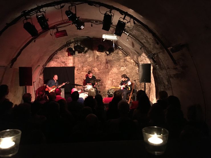 In einem gewölbten, steinernen Raum befinden sich drei Leute auf einer Bühne und machen Musik. Dahinter ist ein Publikum mit vielen Menschen zu sehen. 