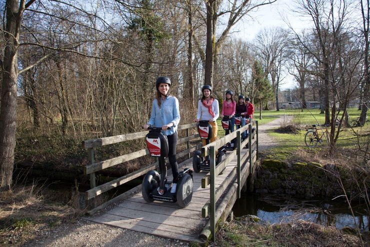 Eine Gruppe von 5 Menschen mit Helmen, die gerade auf Segways über eine Holzbrücke, über einen Bach fahren. In der Umgebung hat es viel Gras, Sträucher und Bäume. 