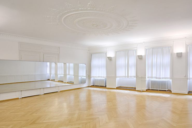 Une grande salle aux murs blancs et sol en plancher. Il y a un grand miroir sur le mur et un beau stuc au plafond. Il y a aussi beaucoup de lumière du jour par les grandes fenêtres.