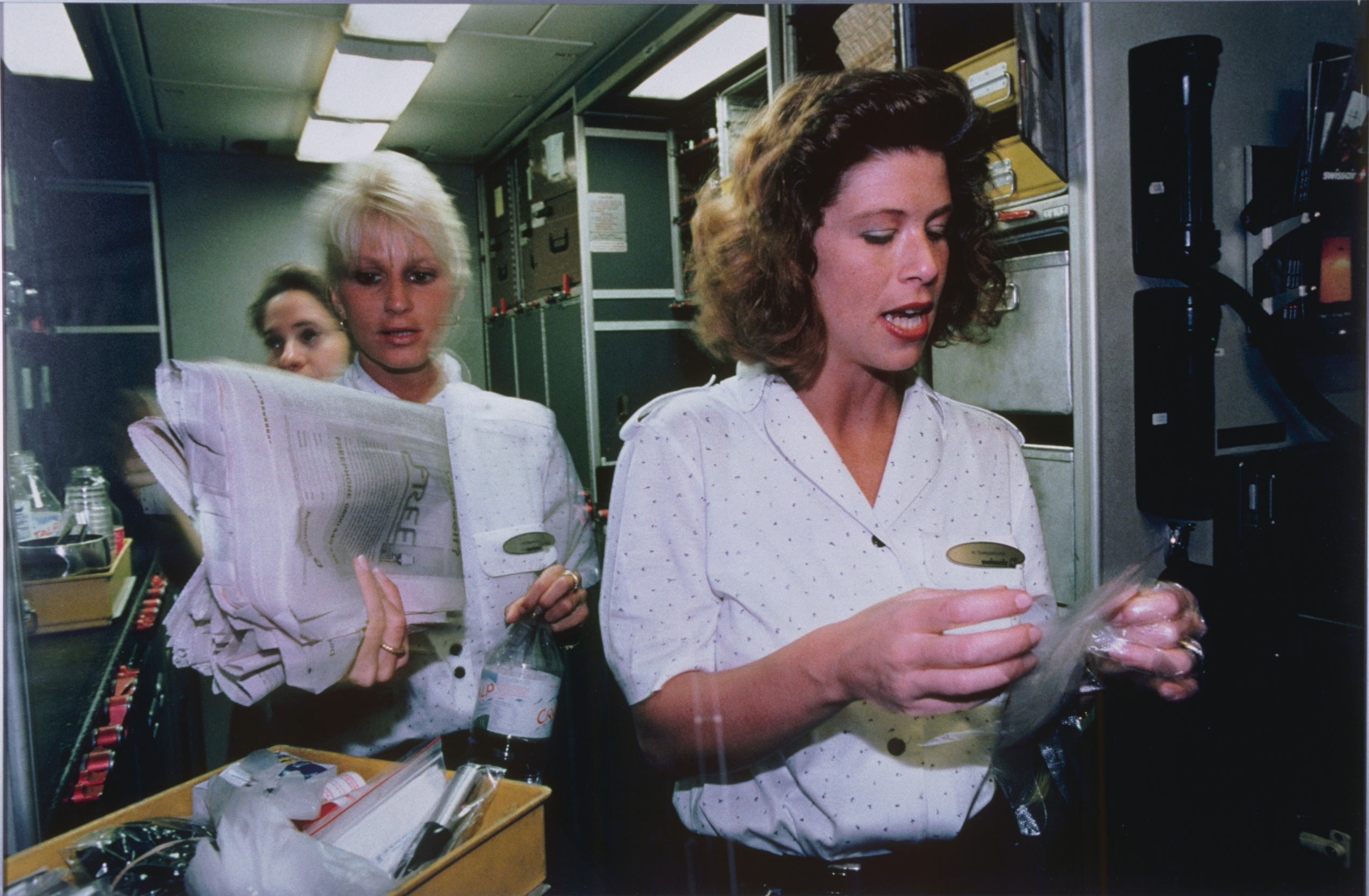 La photo montre trois femmes avec des chemisiers blancs. Une femme tient un journal et une bouteille d'eau et une autre tient quelque chose enveloppé dans du plastique. EIles portent également un badge doré.