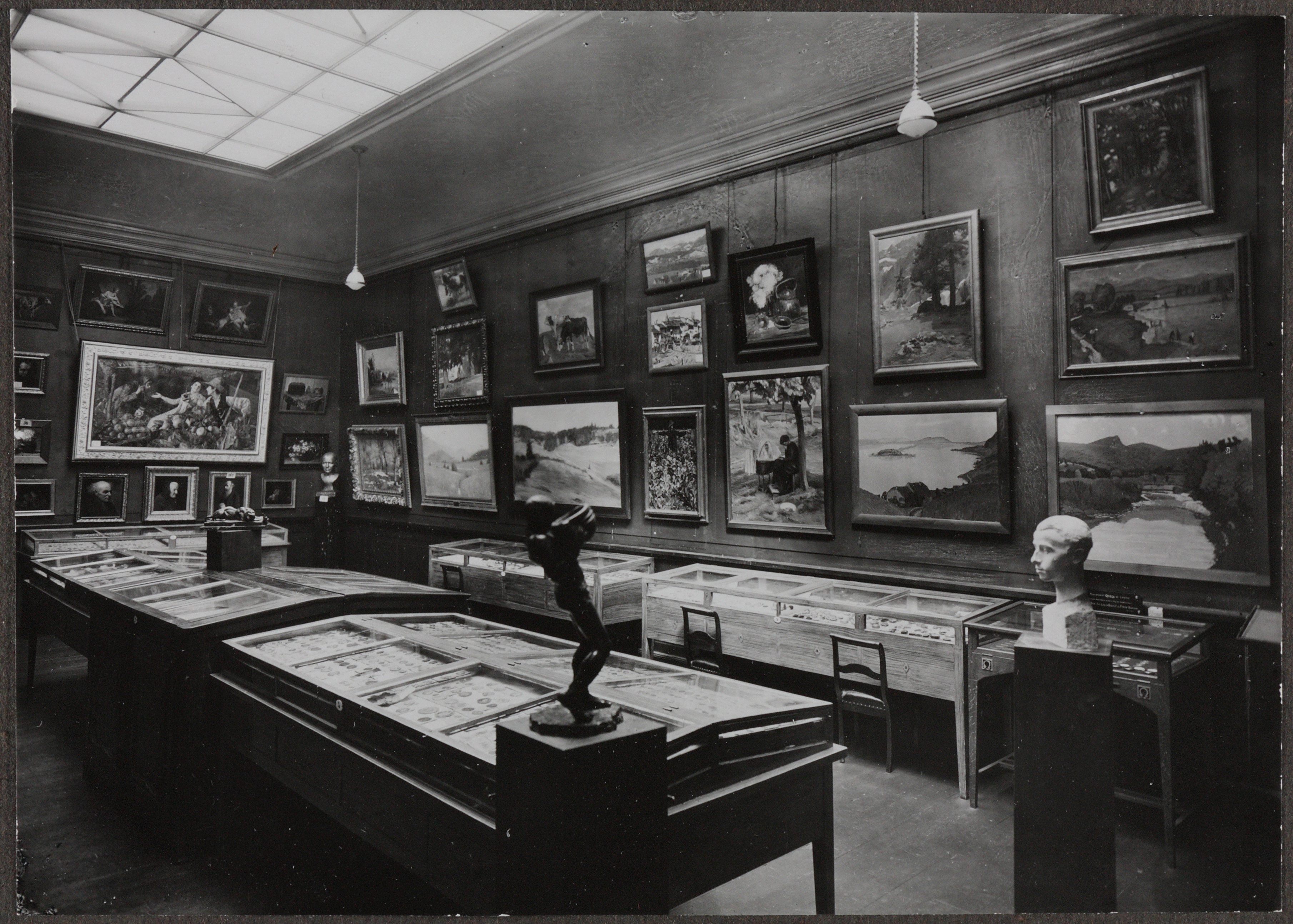 Photographie en noir et blanc d'une salle avec de nombreux tableaux, sculptures et vitrines avec des chaises à l'avant. La lumière passe par une fenêtre au plafond.