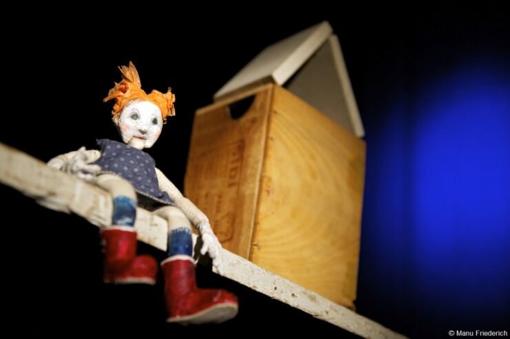 Eine kleine Puppe mit orangen Haaren, blauem Kleid und roten Stiefeln sitzt auf einem Holzbrett. Daneben steht eine Holzkiste, sonst ist das Bild schwarz.