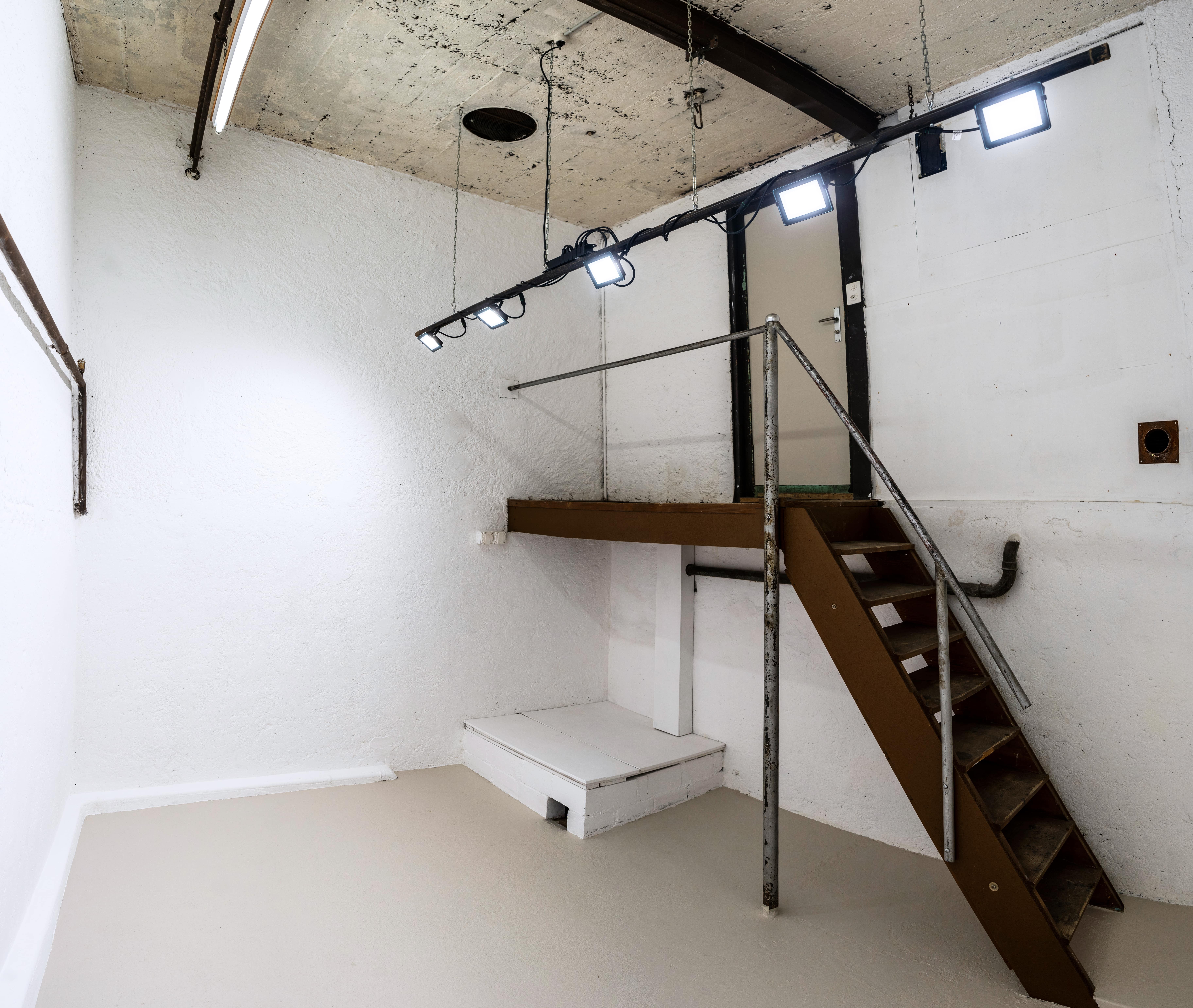 Ein hoher weisser Raum mit hellem Licht, einer Treppe und verschiedenen Eisenrohren. 
