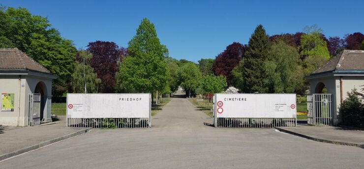 Portail du cimetière principal de Madretsch avec une maisonnette de portier de chaque côté de l'entrée. On voit en arrière-plan la route asphaltée avec une allée d'arbres qui s'avance dans le cimetière.